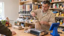 Comerciante de un almacén de barrio vaciando un cucharón de lentejas dentro de una bolsa de papel que sostiene abierta, con la vista puesta en el grano; lleva cotona beige de manga larga. Sobre el mesón de madera, la balanza, un ovillo de hilo y las bolsas de papel dobladas, y a un costado el terminal del punto de venta de espaldas; al fondo, los dispensadores de granel y las repisas con conservas. En primer plano y desenfocados, el hombro y la mano de quien está comprando, junto al canasto con verduras.