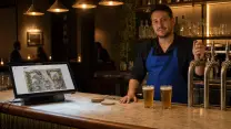 Barman tras la barra de mármol de un pub de noche, con la mano en el tirador de cerveza y el terminal del punto de venta mostrando el plano del salón; al fondo, mesas ocupadas.