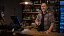 Comerciante de una botillería de noche, con camisa a cuadros remangada, dejando una botella sobre el mesón de madera y con la otra mano apoyada; a un costado el terminal del punto de venta de espaldas, y detrás el refrigerador iluminado y las repisas con botellas, cajas de cartón y carteles de precio escritos a mano.
