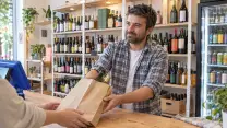 Comerciante de una botillería entregando por el mesón una bolsa de papel con botellas, con la vista puesta en el cliente, del que sólo se ven las manos; lleva camisa a cuadros remangada sobre polera blanca. Detrás, las repisas con botellas de distintos formatos y colores y el refrigerador de bebidas; a un costado, el terminal del punto de venta de espaldas.