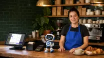 Barista de una cafetería sonriendo tras su mostrador junto a la mascota robot de TransaK; en el mesón, el terminal del punto de venta con la pantalla de cobro, el lector de tarjetas y la impresora de boletas.