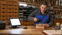 Ferretero pasando una caja de pernos por el lector de códigos tras el mesón de madera de su local; a su lado, el terminal del punto de venta con el control de existencias en pantalla y la impresora, y al fondo la muralla de cajones y las bandejas de tornillos.