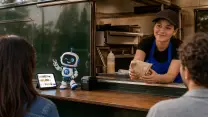 Trabajadora de un food truck asomada a la ventanilla entregando un pedido envuelto en papel; sobre el mesón, la tablet del punto de venta con el catálogo en pantalla, la impresora de boletas y la figurina de la mascota de TransaK, con la cocina de acero al fondo.