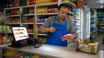 Comerciante de un minimarket pasando un producto por el lector de códigos tras el mesón de la caja; a su lado, el terminal del punto de venta con el catálogo en pantalla, la impresora de boletas y el lector de tarjetas, con las góndolas y el refrigerador detrás.