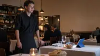 Garzón de un restaurante, con delantal negro largo, de pie junto a una mesa vestida y con una mano apoyada en el respaldo de una silla; sobre el mantel, la tablet del punto de venta en su soporte, de espaldas a la cámara; al fondo, la barra y otras mesas ocupadas.
